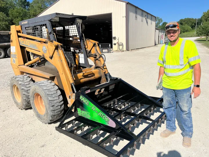 B-Built Rake-N-Ator (78 Zoll) an einem Case-Kompaktlader, daneben ein Mann in Warnweste